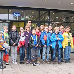 Eine Gruppe von Kindern und zwei Erwachsenen stehen vor einem Gebäude mit Glasfassade